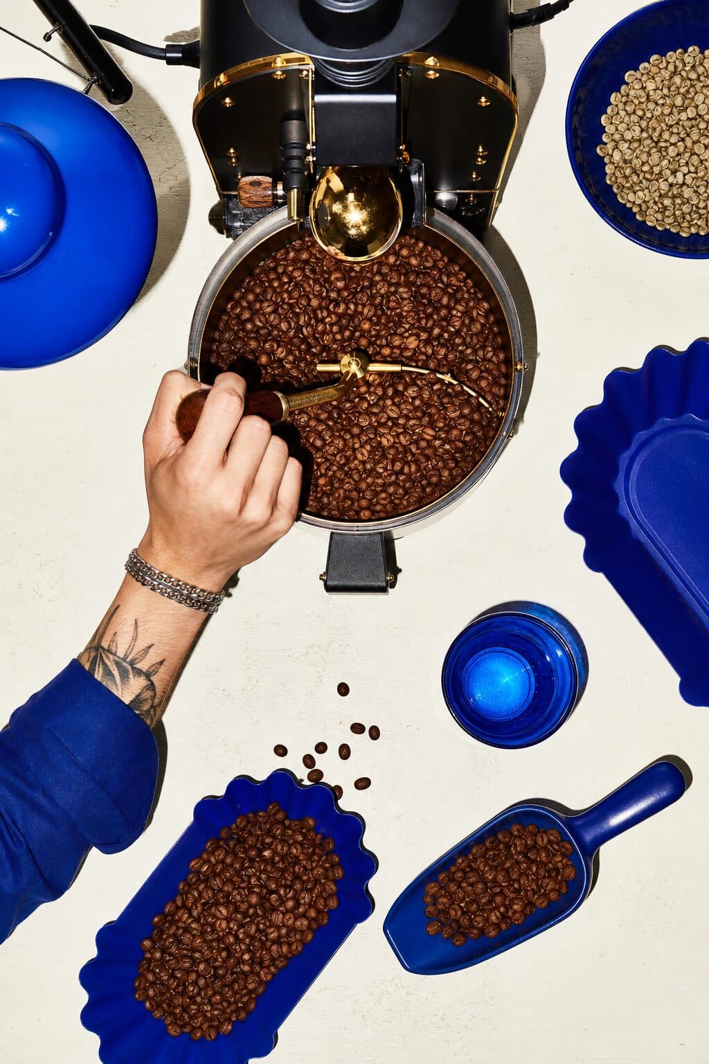 Straight down over the roaster: a hand drawing a scoop of beans from the drum, blue trays and a bowl of green beans around it