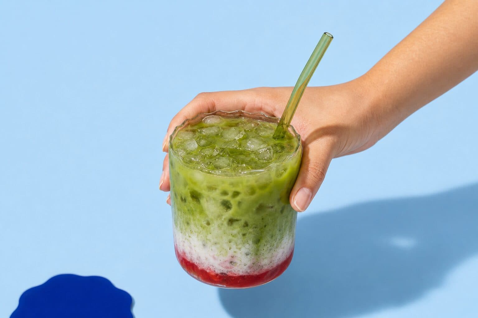 A strawberry matcha held up in a rippled glass, layered green over white over red, against pale blue