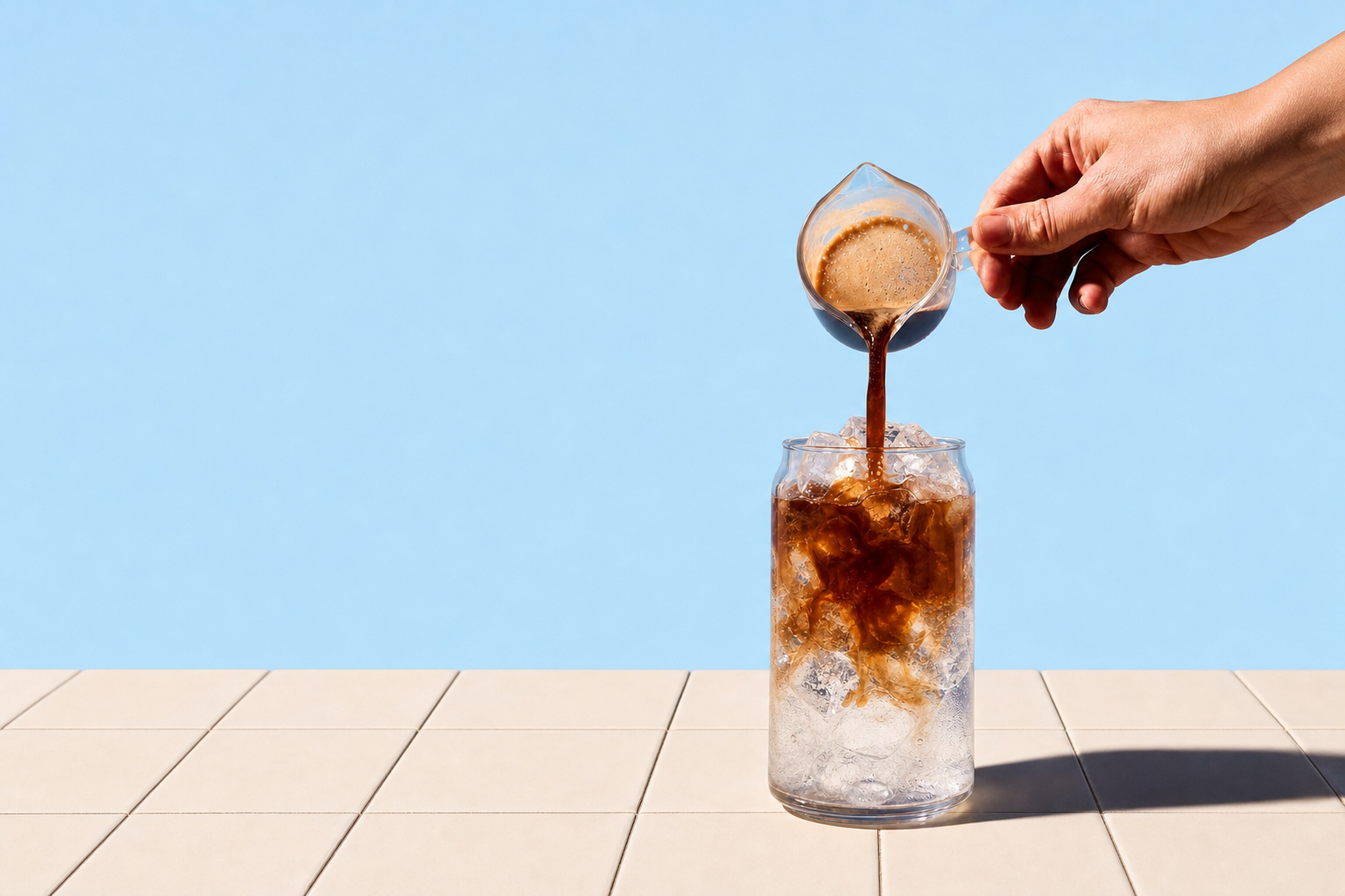 Espresso poured from a glass jug into a tall glass of ice on a cream tiled counter, a pale sky-blue wall behind